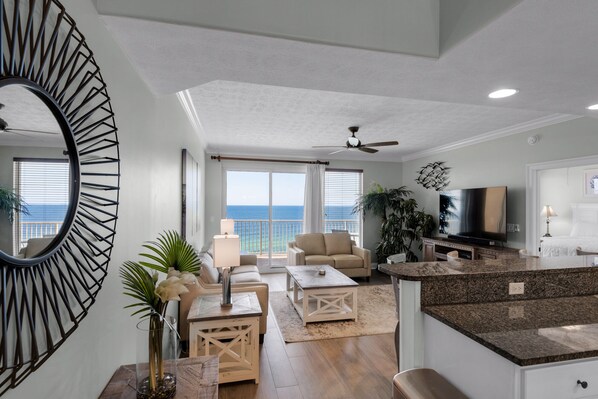 A spacious living area with a large window overlooking the ocean, a ceiling fan, and a modern kitchen with granite countertops.