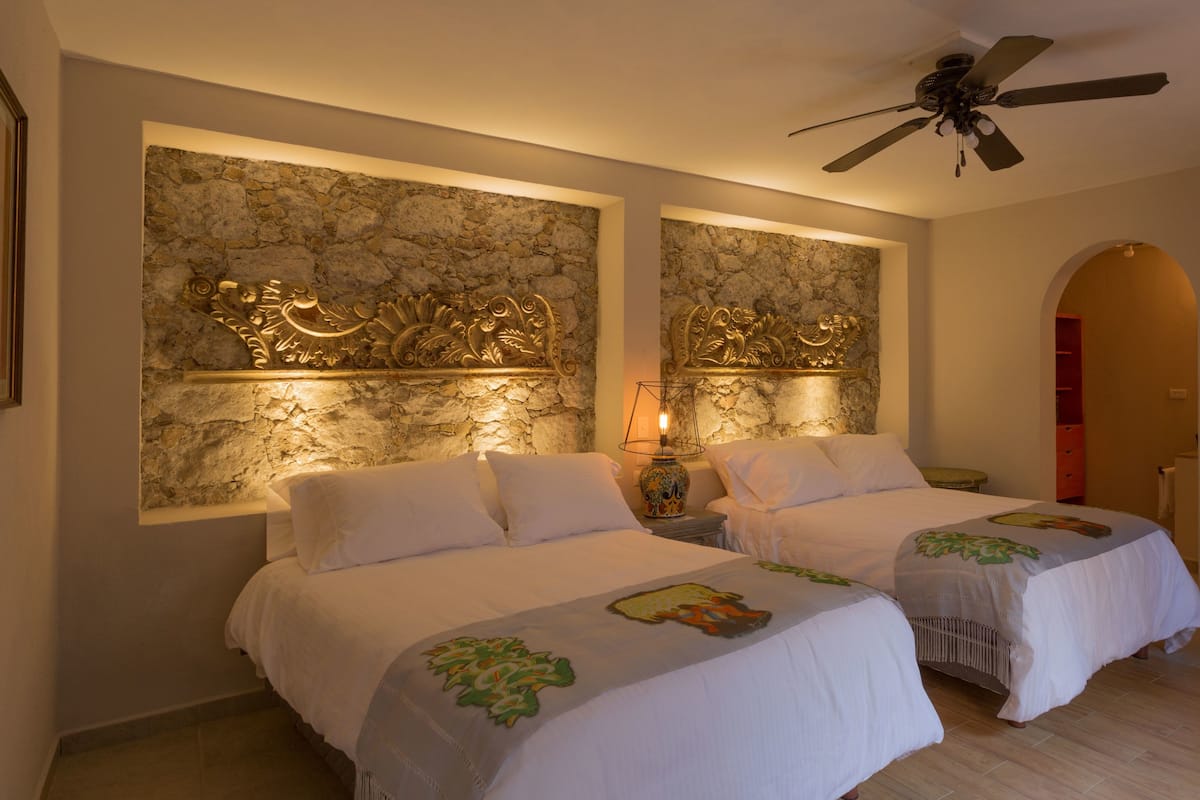 A hotel room with two beds, a stone wall, a ceiling fan, and a nightstand with a lamp.