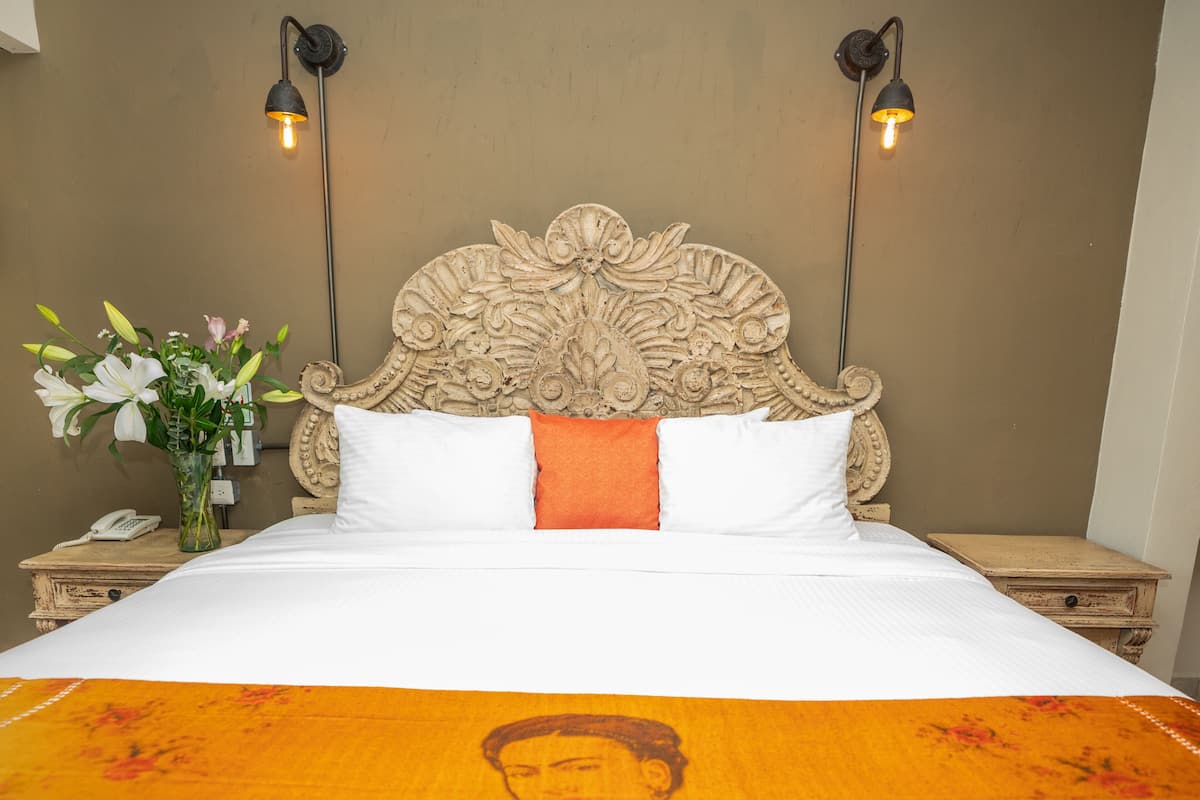 A bed with a white and orange bedspread, a wooden headboard with intricate carvings, two bedside lamps, a nightstand with a phone, and a vase with flowers.
