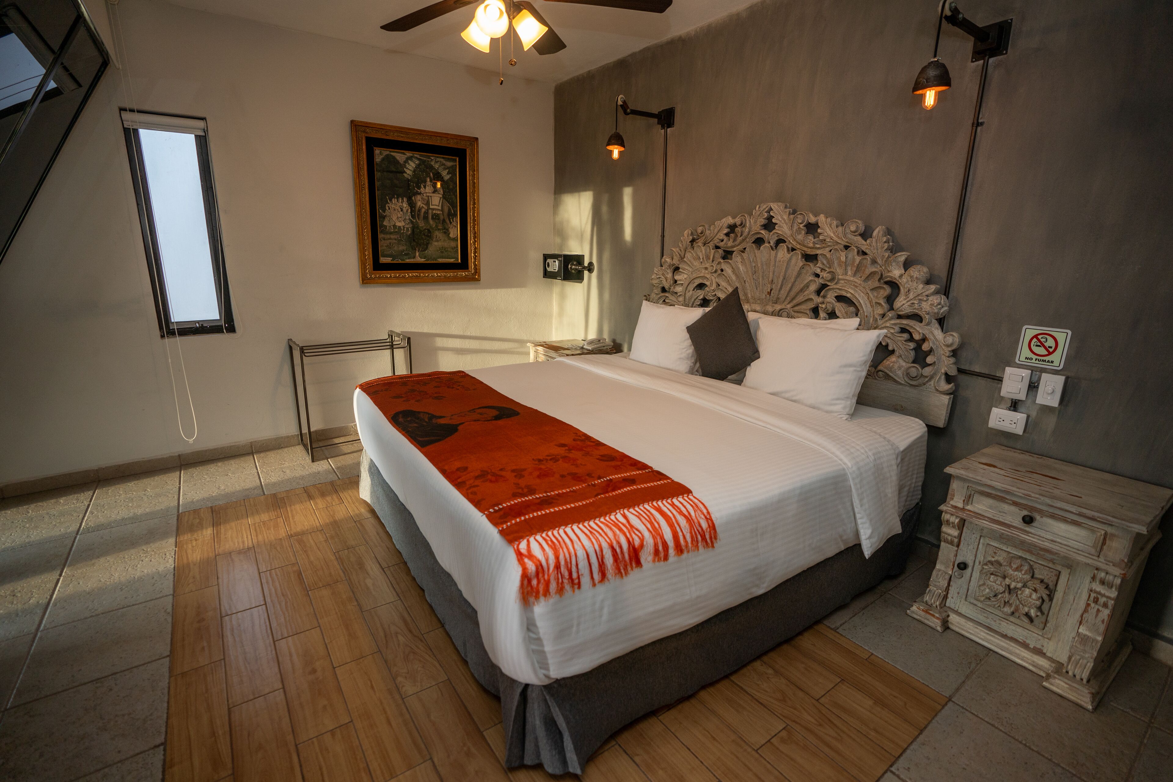 A bedroom with a large bed, a painting on the wall, a ceiling fan, and a nightstand with a lamp.