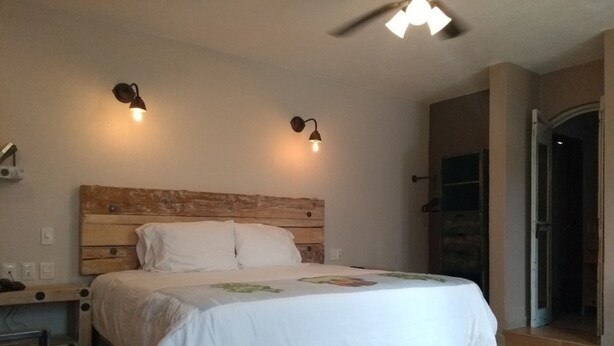 Quarto com cabeceira de madeira, duas luminárias de cabeceira, cama com colcha branca, uma mesinha e uma cadeira.