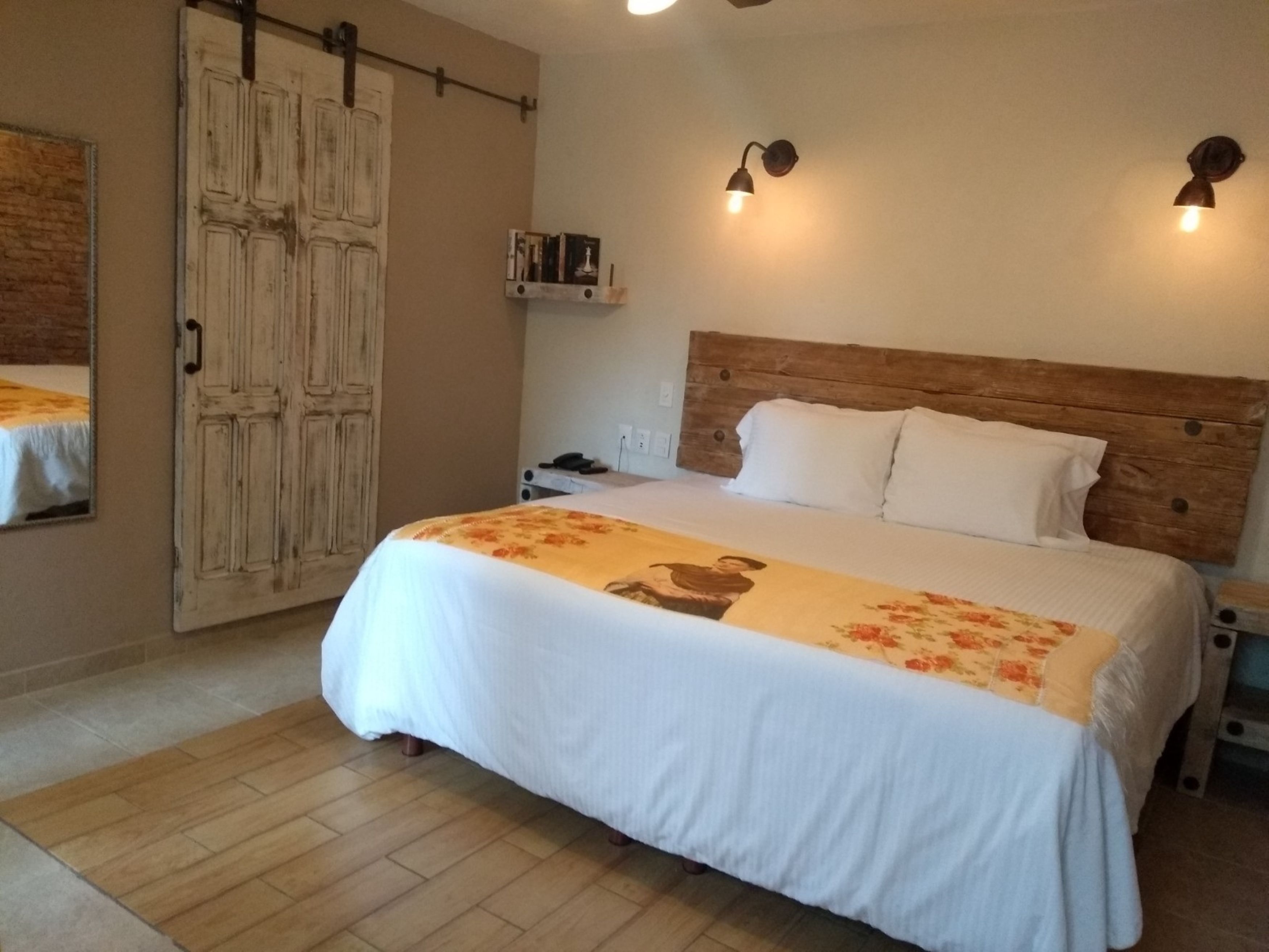 Une chambre à coucher avec une tête de lit en bois, un lit avec une literie blanche, deux lampes fixées au mur et une porte en bois.