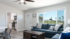 A modern living room with a dark wooden coffee table, a blue sofa with patterned cushions, and a view of a golf course through large windows. - 1417G - "After Dune Delight" (Ventura)