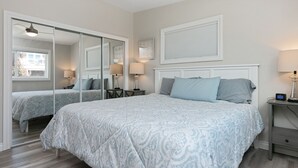 A bedroom with a large bed, a nightstand, a mirror, and a window with blinds. - 1417G - "After Dune Delight" (Ventura)