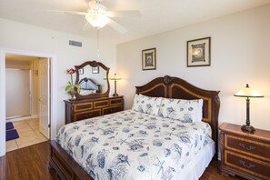 A bedroom with a large bed, a wooden dresser, a nightstand, a ceiling fan, and two framed pictures on the wall.