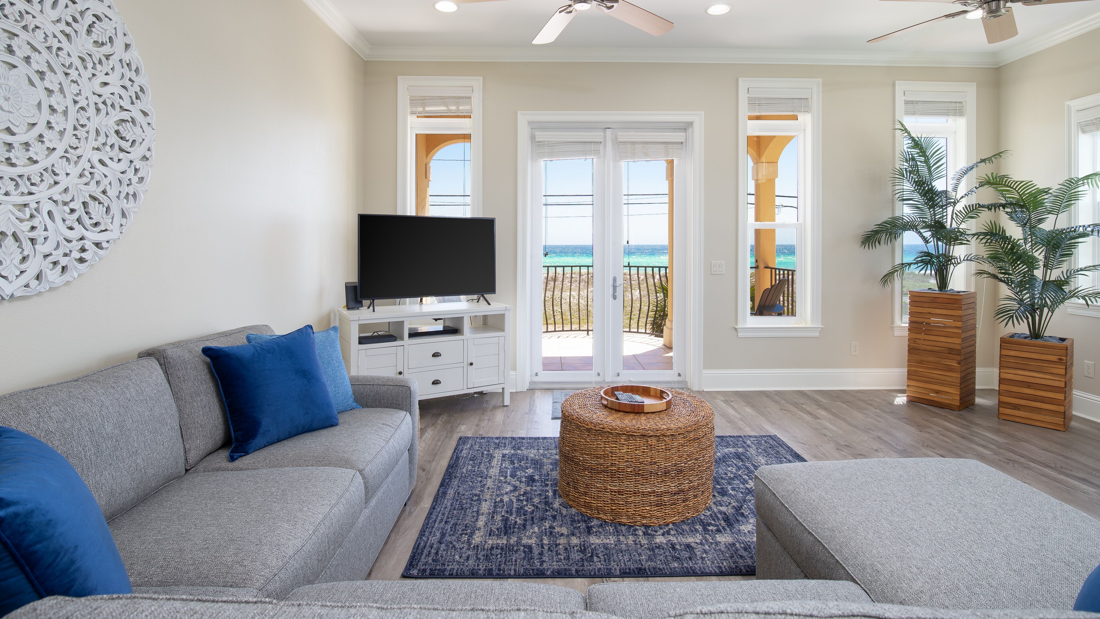 A living room with a grey sofa, a flat-screen TV, a woven ottoman, and a view of the ocean through large sliding doors.