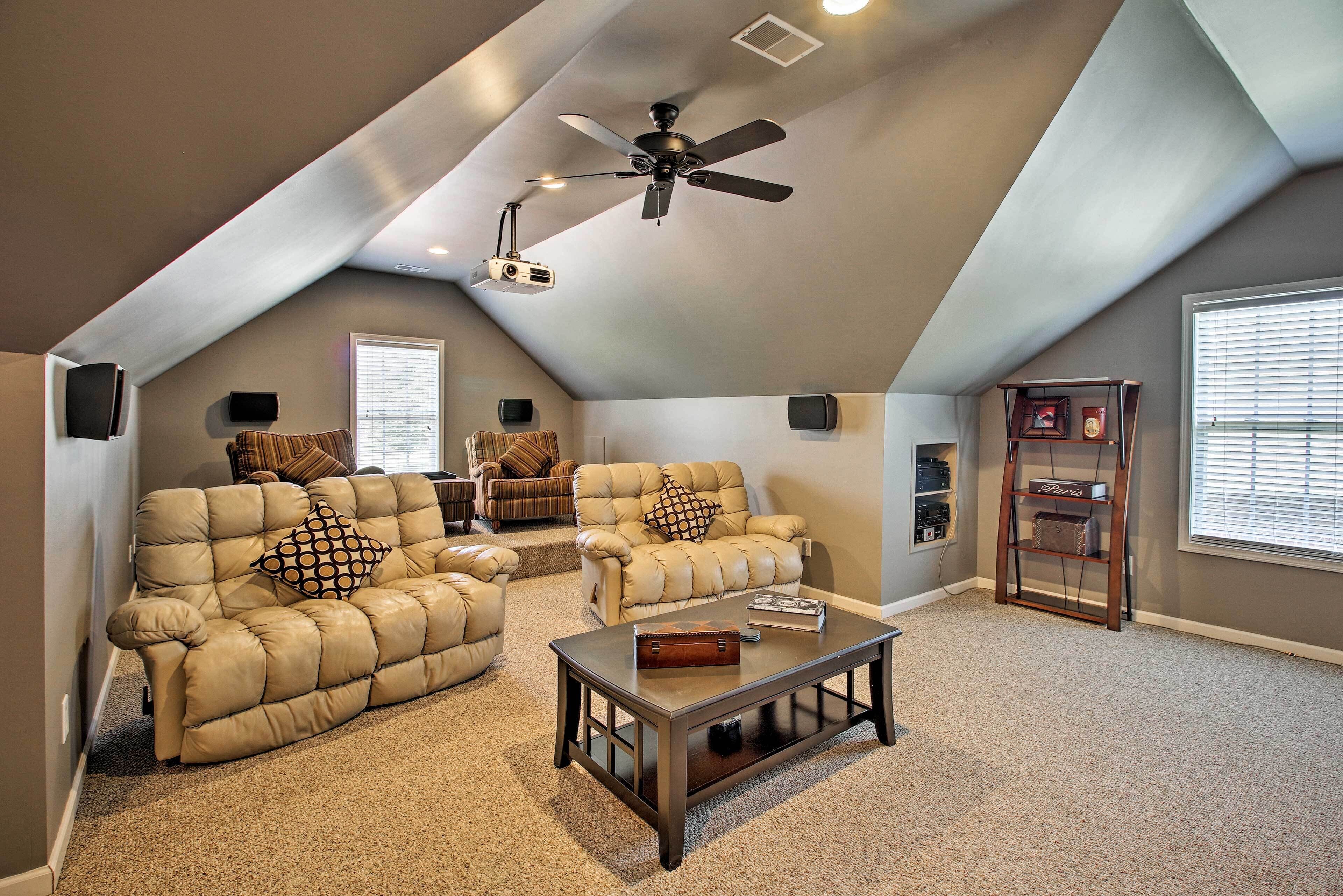 A spacious living room with a high ceiling, a ceiling fan, and a projector. There are two leather sofas with patterned cushions, a wooden coffee table, and a bookshelf.