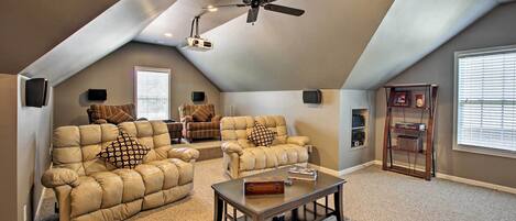 A spacious living room with a high ceiling, a ceiling fan, and a projector. There are two leather sofas with patterned cushions, a wooden coffee table, and a bookshelf.
