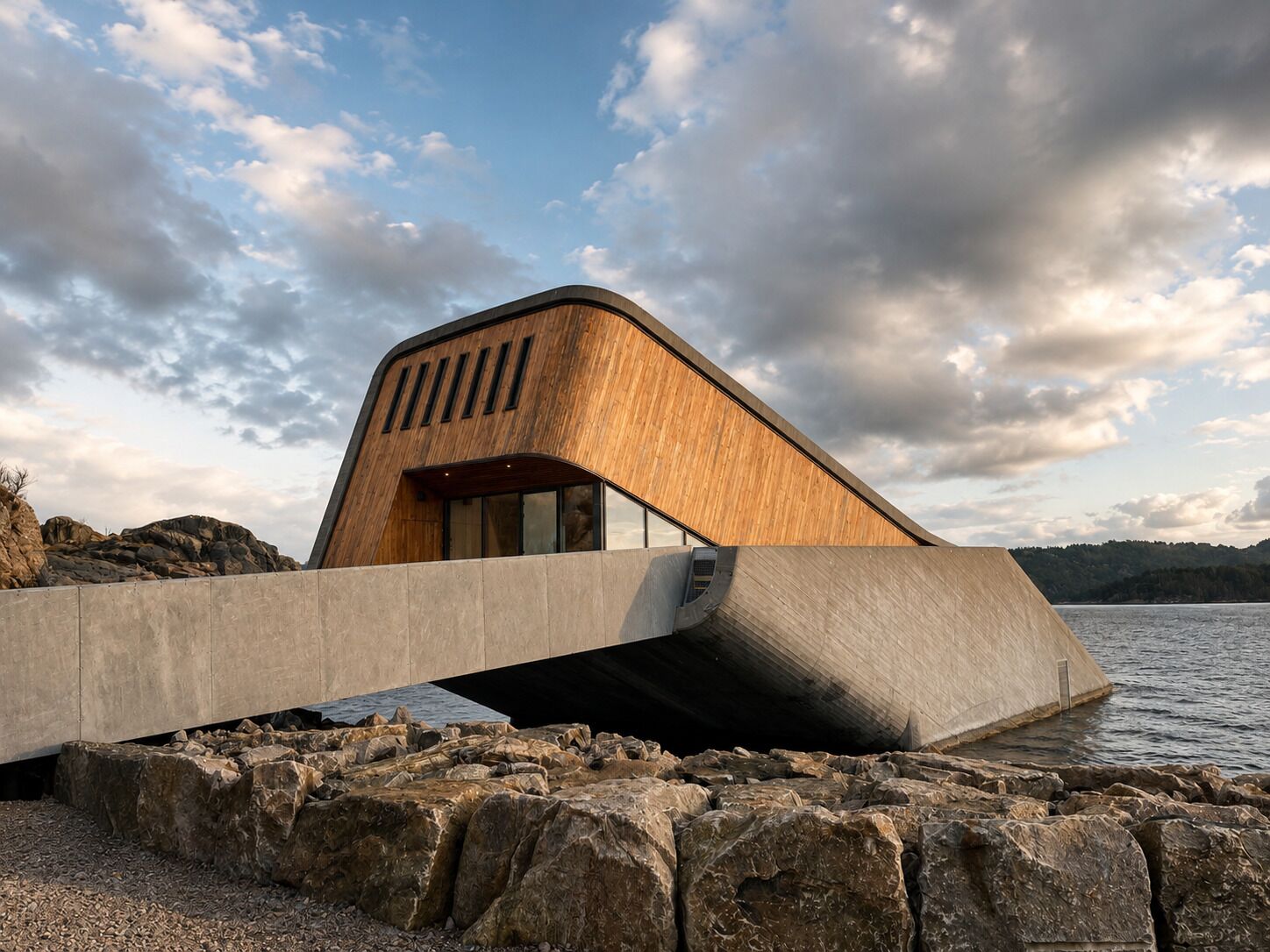 Un edificio moderno con facciata in legno, un ponte in cemento e un litorale roccioso.