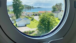 Une vue à travers une fenêtre sur un quartier résidentiel, avec des maisons, une pelouse verdoyante et une étendue d’eau au loin.
