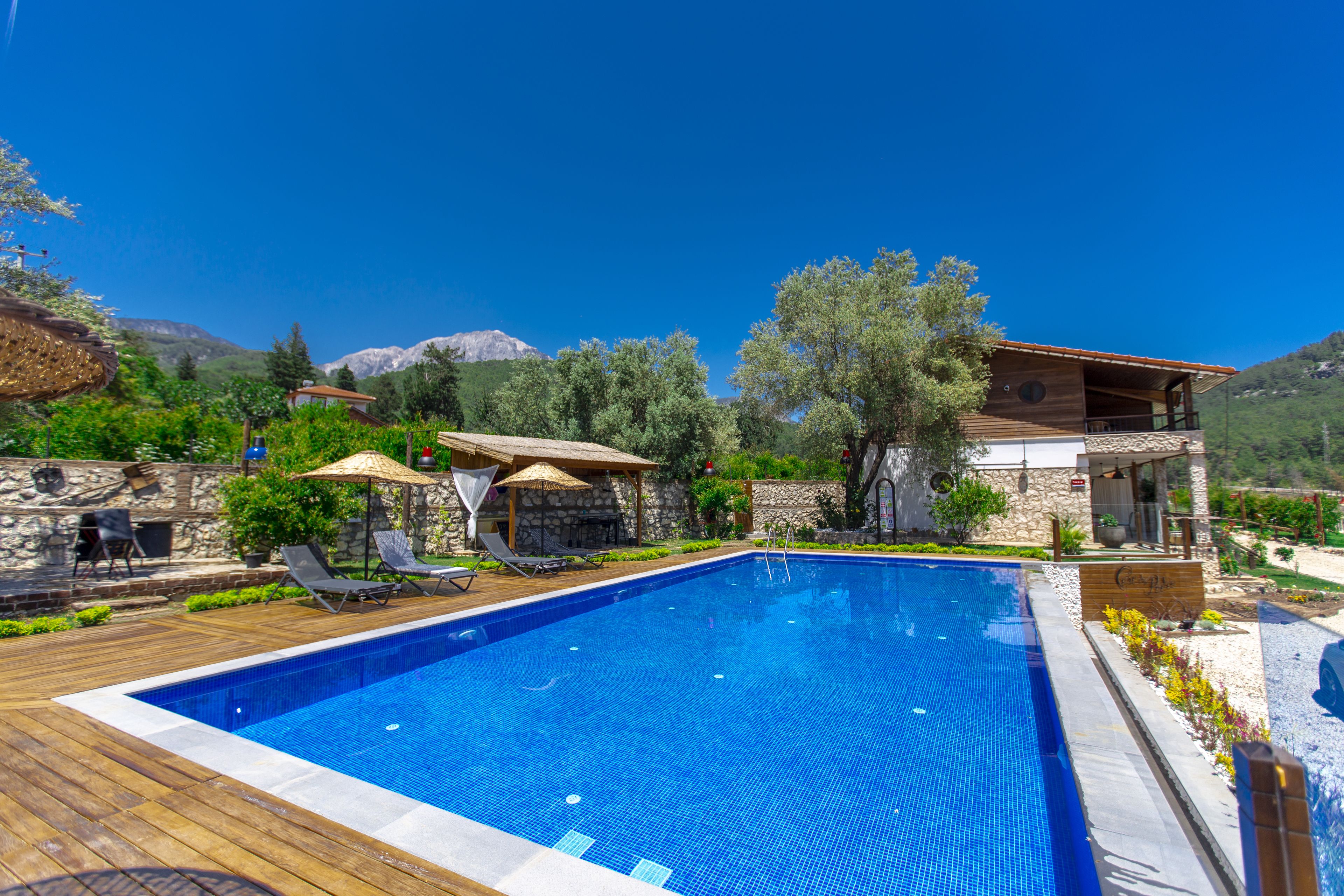 Uma piscina com espreguiçadeiras e guarda-sóis, cercada por paredes de pedra e vegetação, com montanhas ao fundo.