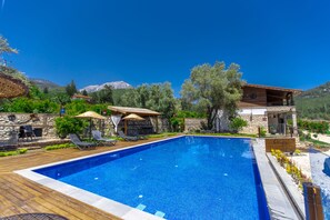 A swimming pool with lounge chairs and umbrellas, surrounded by stone walls and greenery, with mountains in the background.