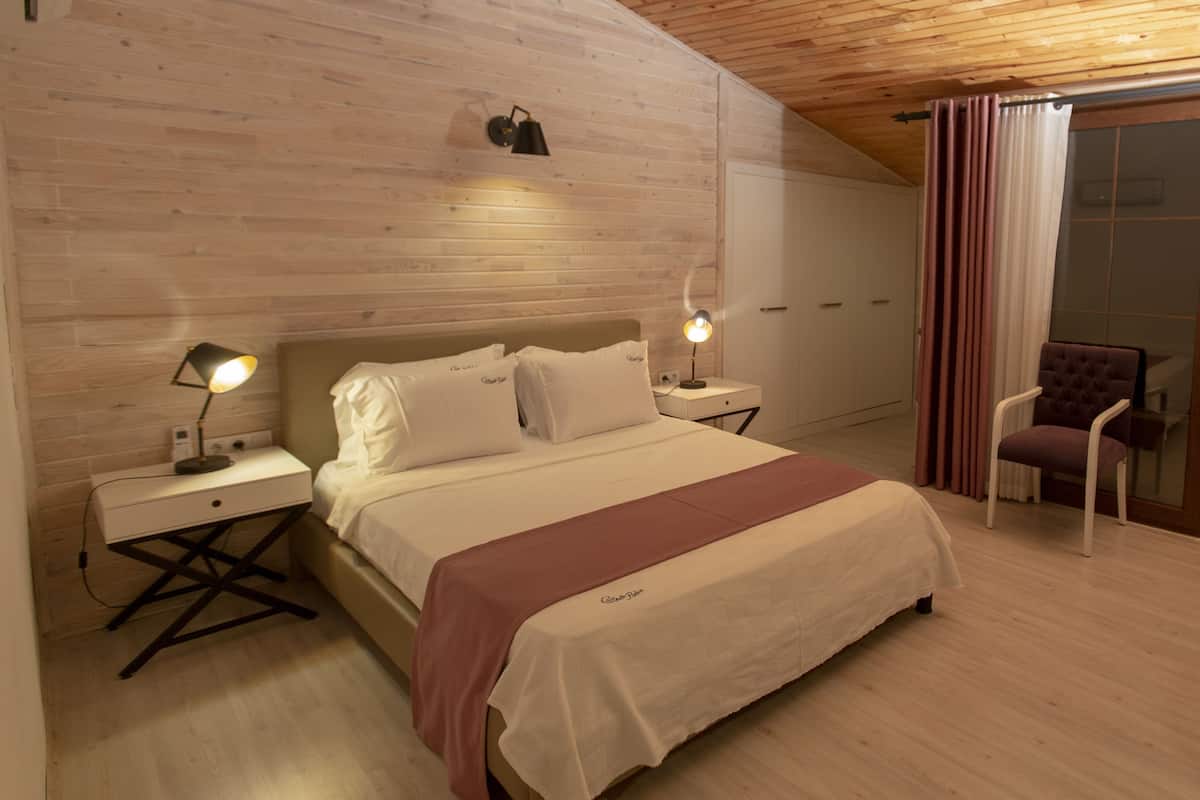 Ein Schlafzimmer mit Holzbalkendecke, einem Bett mit weißer Bettwäsche, zwei Nachttischlampen, einem Nachttisch, einem Sessel und einer Glastür mit Vorhängen.