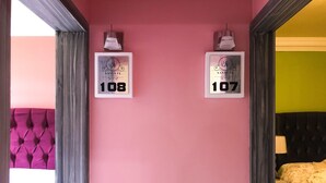 Habitación de hotel con pared rosa, dos letreros enmarcados con los números de habitación 108 y 107, cabecera capitonada y cama con ropa de cama floral.