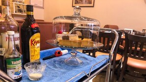 Un carrito de servicio con un pastel sobre un soporte de vidrio, una botella de Kahlúa, una botella de extracto de vainilla y un pequeño cuenco de vidrio con azúcar.