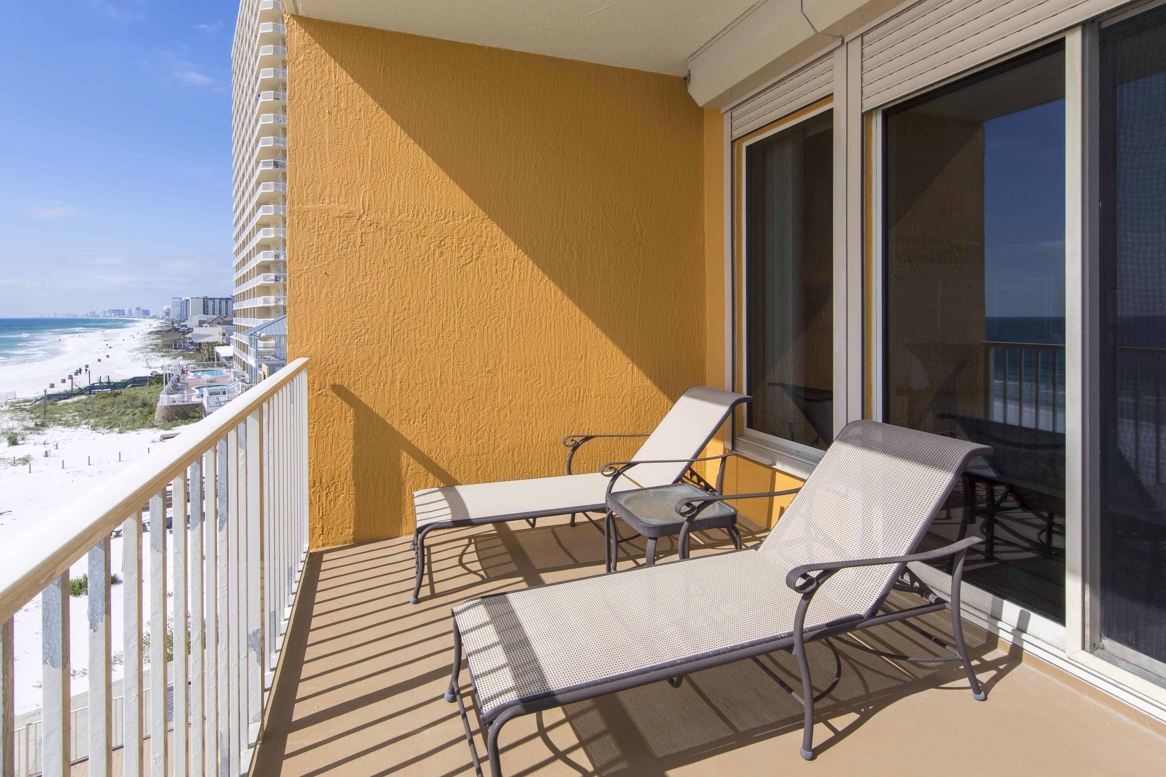 A balcony with two lounge chairs and a small table, overlooking a beach with buildings in the background.