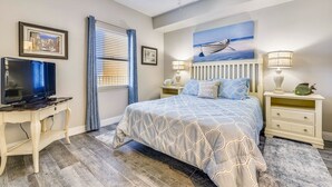 Een slaapkamer met een bed, een televisie op een bureau, een plafondventilator en een schilderij van een boot aan de muur.