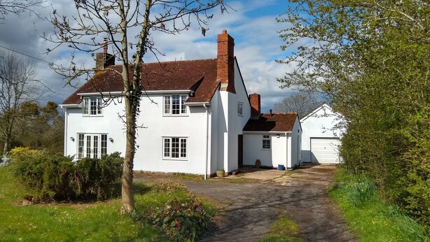 Une maison blanche à deux étages, avec un toit recouvert de tuiles rouges, une cheminée et un petit garage attenant.