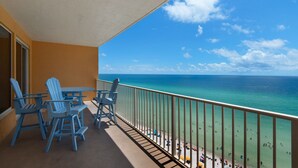 A balcony with blue chairs and a table, overlooking a beach with people and umbrellas. - 1107-11th Floor-BEACH CHAIRS & UMBRELLA INCL-Outdoor Pool-AMAZING BEACH  VIEW! (Panama City Beach)