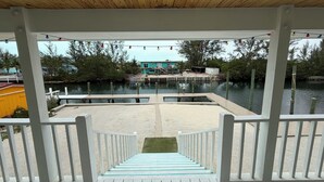 A covered deck with a view of a dock and water, surrounded by greenery and a building in the background.
