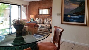 A dining area with a glass table, chairs, a plant, a sofa with a patterned cover, and a painting on the wall.