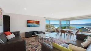 A modern living room with a large window, a flat-screen TV, and a dining area with chairs and a table.