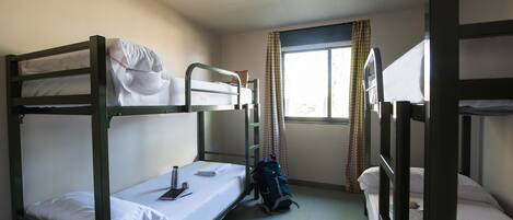 Habitación de residencia con literas, una ventana con cortinas, una mochila en el suelo y una botella de agua sobre la cama.