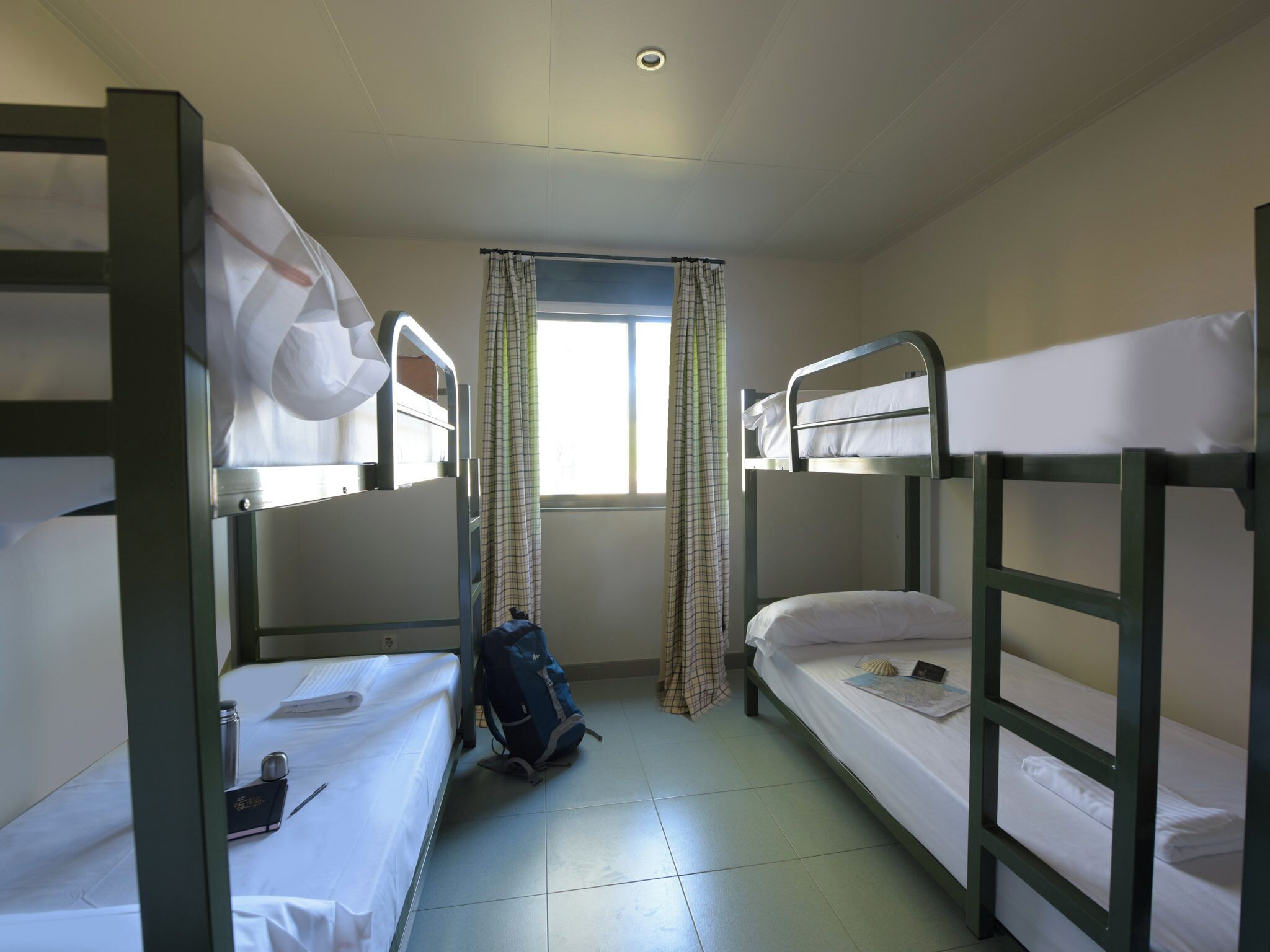 Una habitació de dormitori amb llits superposats, una finestra amb cortines, una motxilla i una ampolla d'aigua a terra.