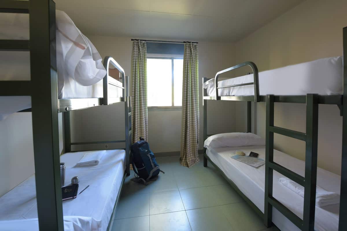 Ein Schlafsaalzimmer mit Etagenbetten, einem Fenster mit Vorhängen, einem Rucksack und einer Wasserflasche auf dem Boden.