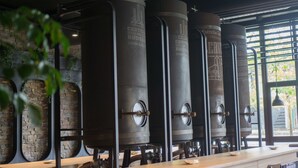 A row of large cylindrical brewing tanks in a modern industrial setting with a wooden counter and stone wall. - Hostel Monte Do Gozo (Santiago de Compostela)