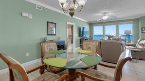 A dining area with a glass table surrounded by wooden chairs with green cushions, a chandelier, and a ceiling fan.