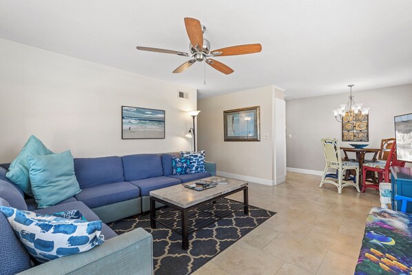 A living room with a blue sofa, a coffee table, a ceiling fan, and a dining area with a chandelier.