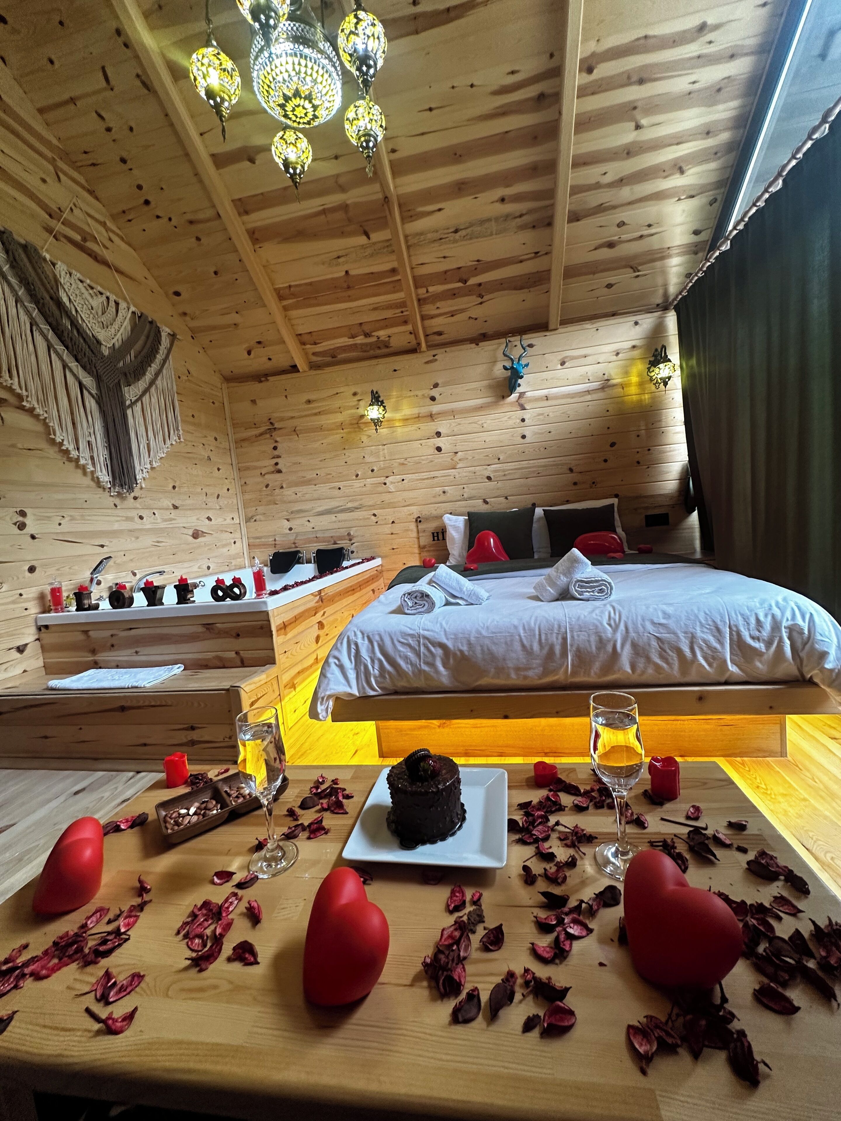 Une pièce chaleureuse, aménagée dans une cabane en bois, avec un lit, une petite table sur laquelle se trouvent une assiette de nourriture et des boissons, et un lustre.