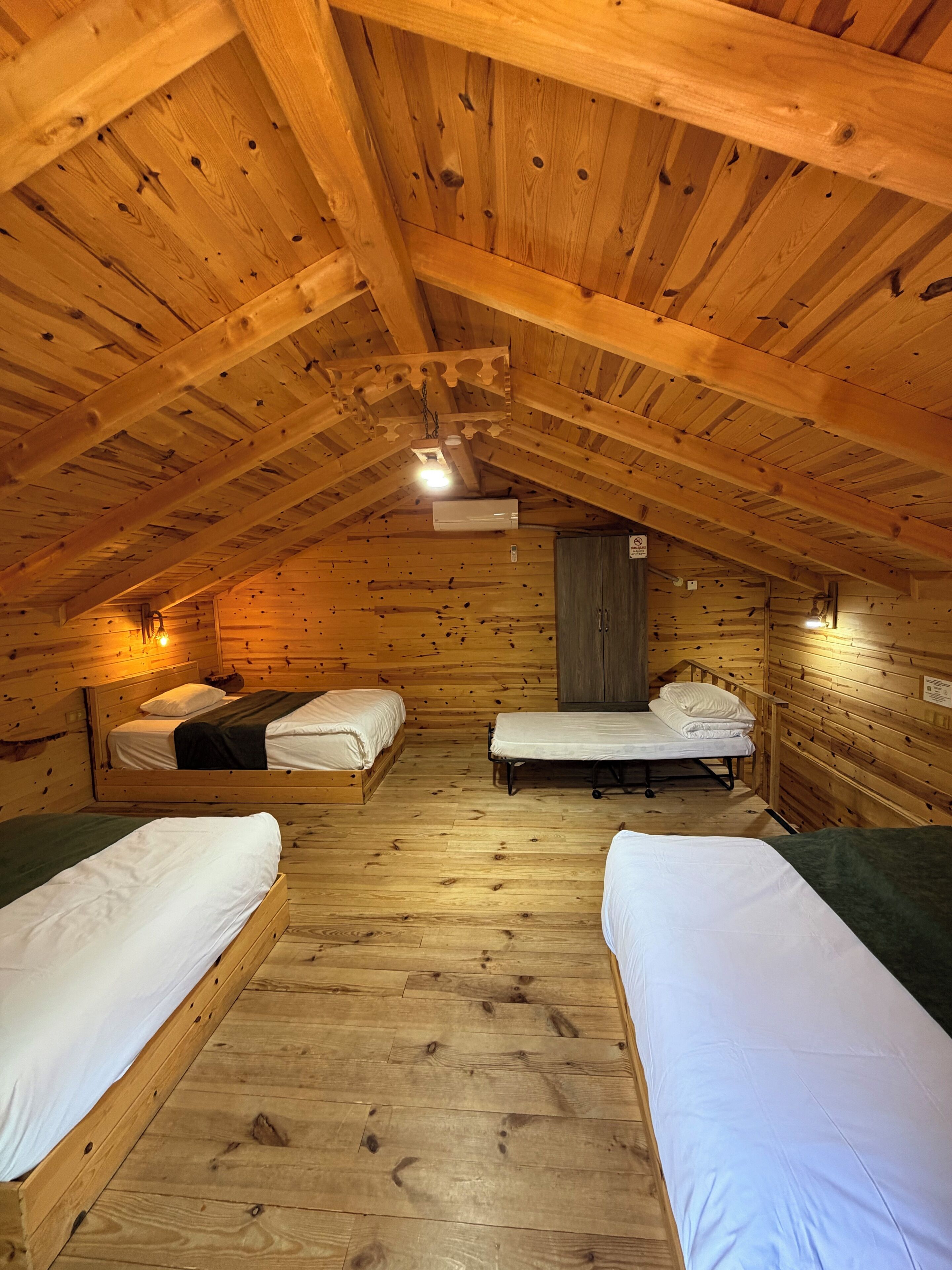 A wooden cabin with four beds arranged in a U-shape, a wooden ceiling with beams, and a single light fixture.