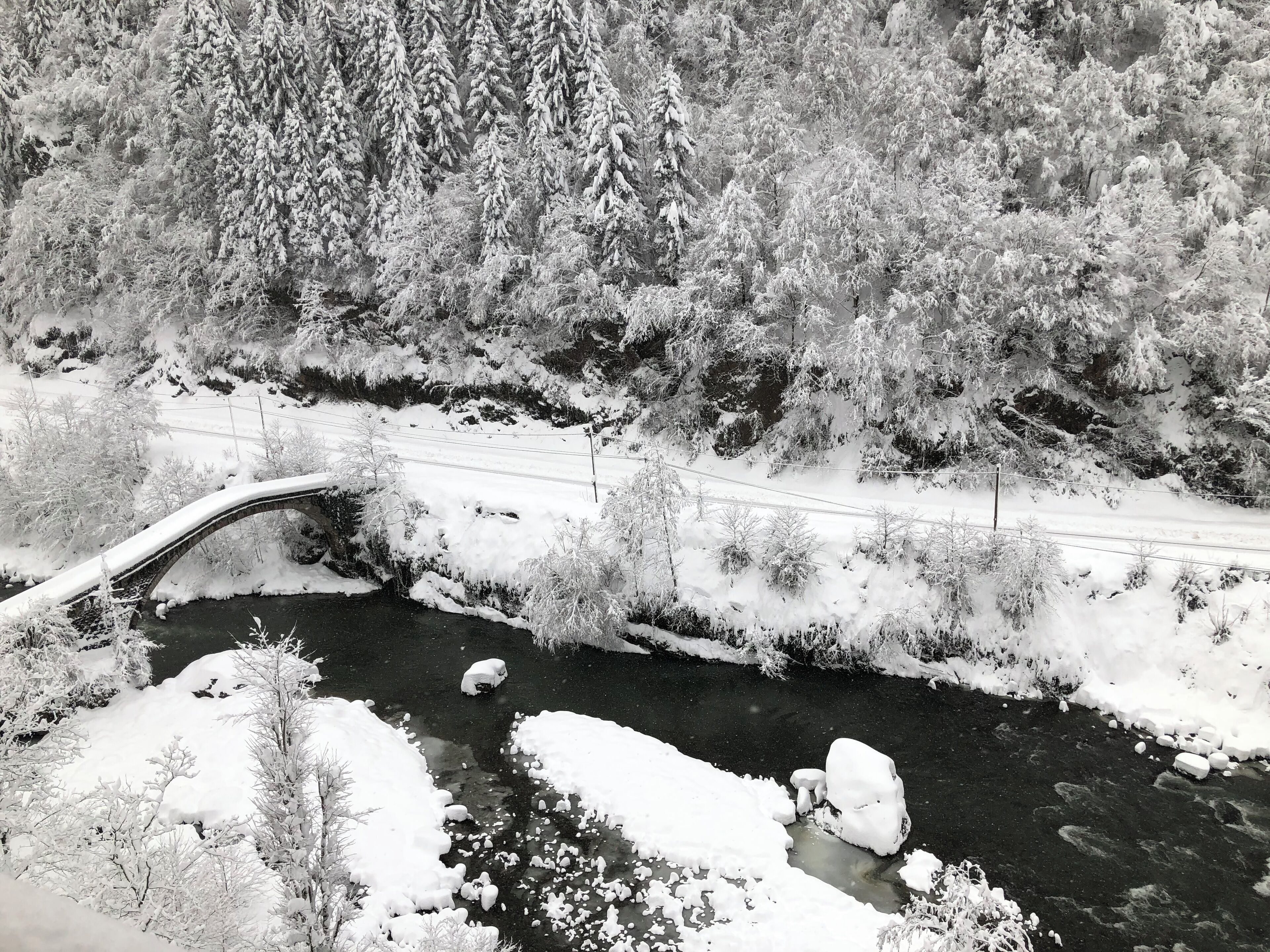 A snow-covered landscape with a bridge over a river, surrounded by trees and a road.