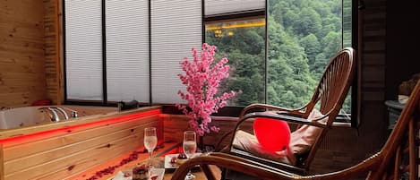 A spa room with a wooden bench, a red exercise ball, a wooden chair, a small table with a glass of water, and a view of a forest through large windows.