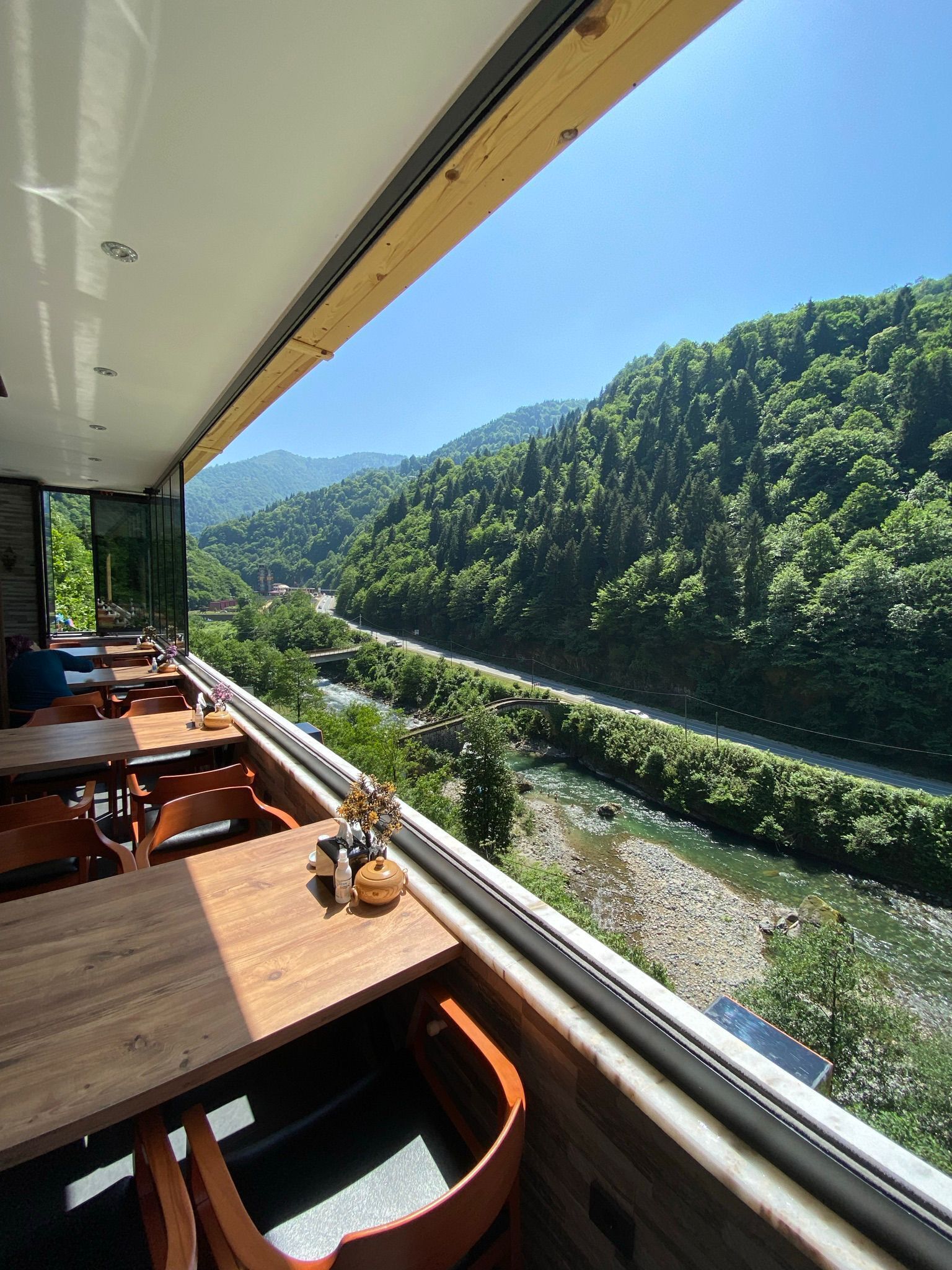 A dining area with wooden tables and chairs, a view of a river, and a forested hillside.