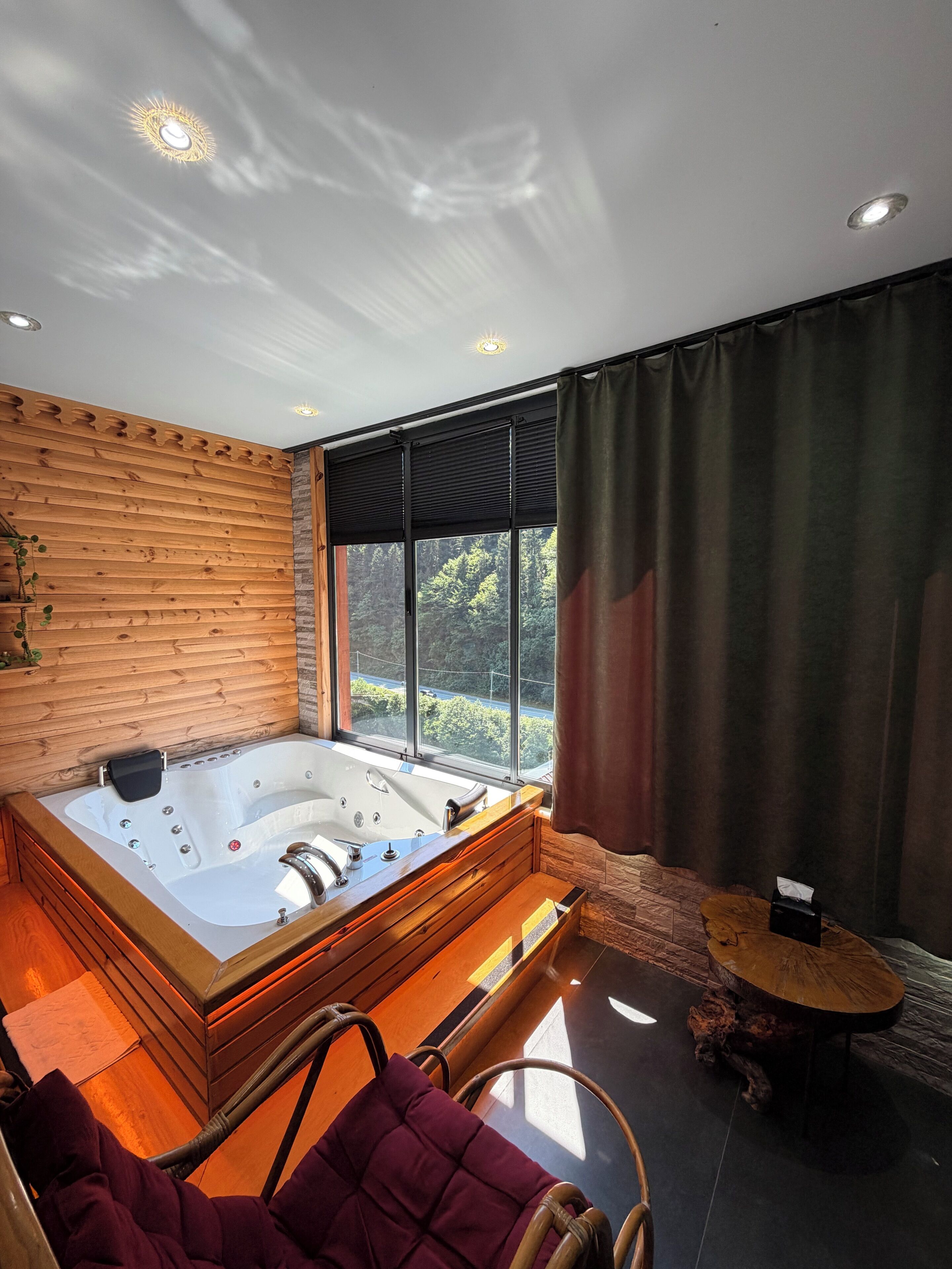 A modern bathroom with a large bathtub, wooden walls, a window with curtains, and a small round table.
