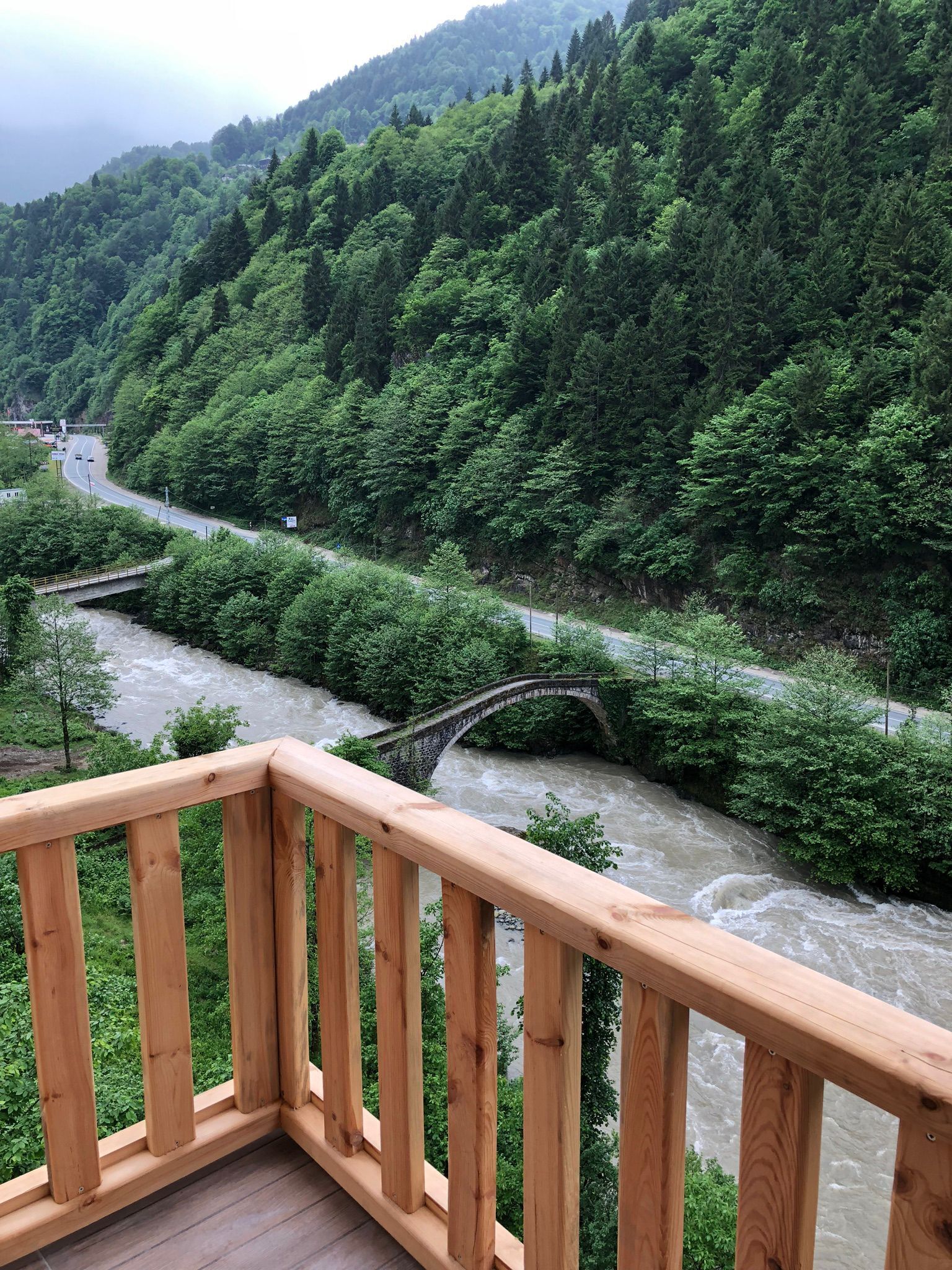A wooden deck overlooking a river with a stone bridge, surrounded by dense green forest.