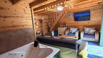 A wooden cabin interior with a large bathtub, two sofas, a small table, and a television.