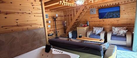 A wooden cabin interior with a large bathtub, two sofas, a small table, and a television.