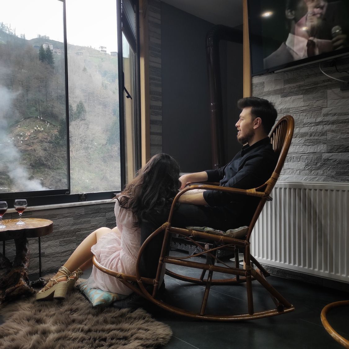 Una habitación con un ventanal grande, una pared de piedra, una silla de madera, una mesita con vasos, y una alfombra de piel.