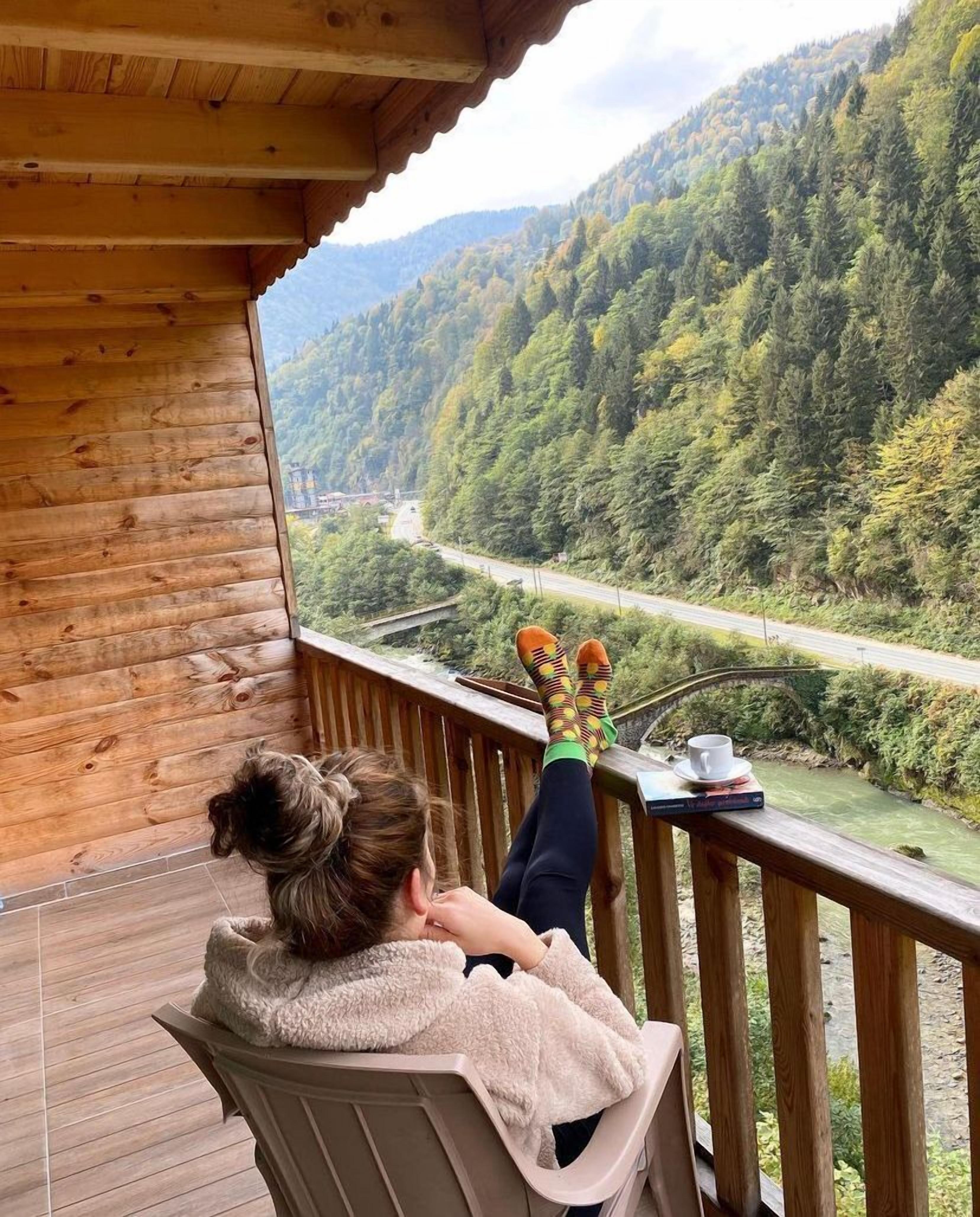 Una persona descansando en una terraza de madera con vista a un valle boscoso y una carretera.