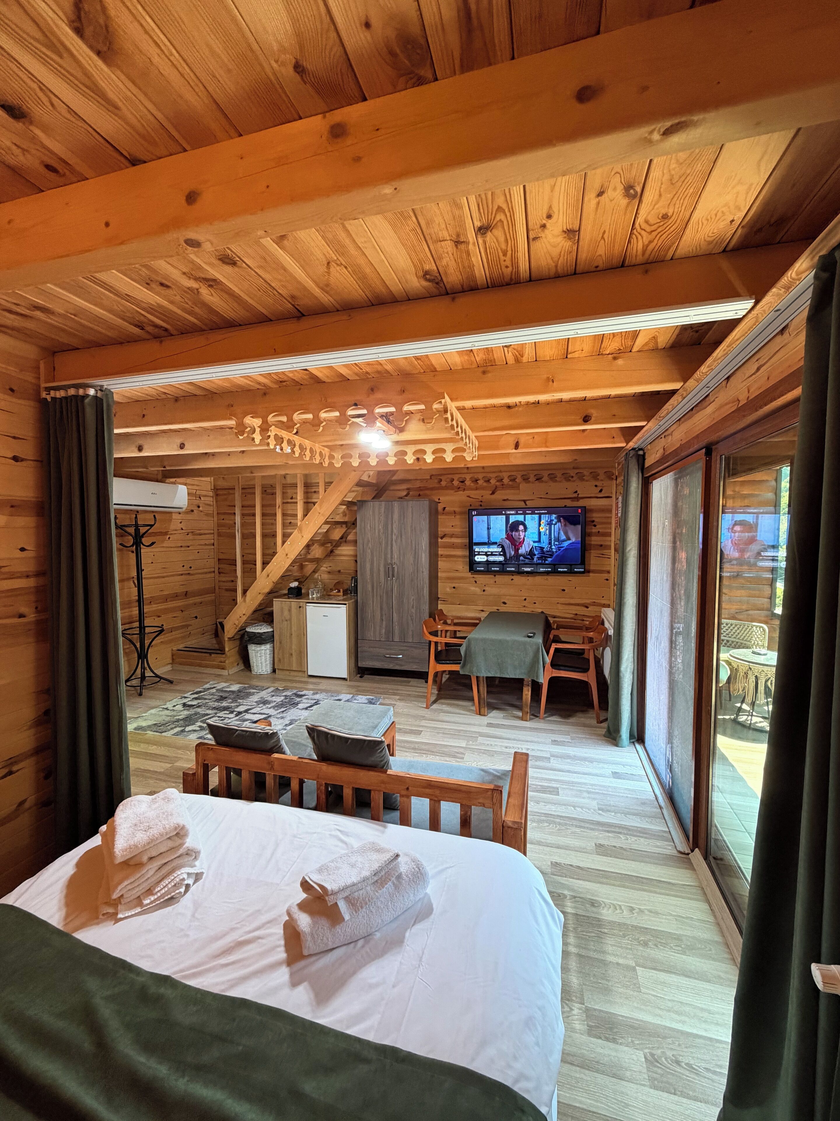 A wooden cabin interior with a bed, a dining area, a TV, and a staircase.
