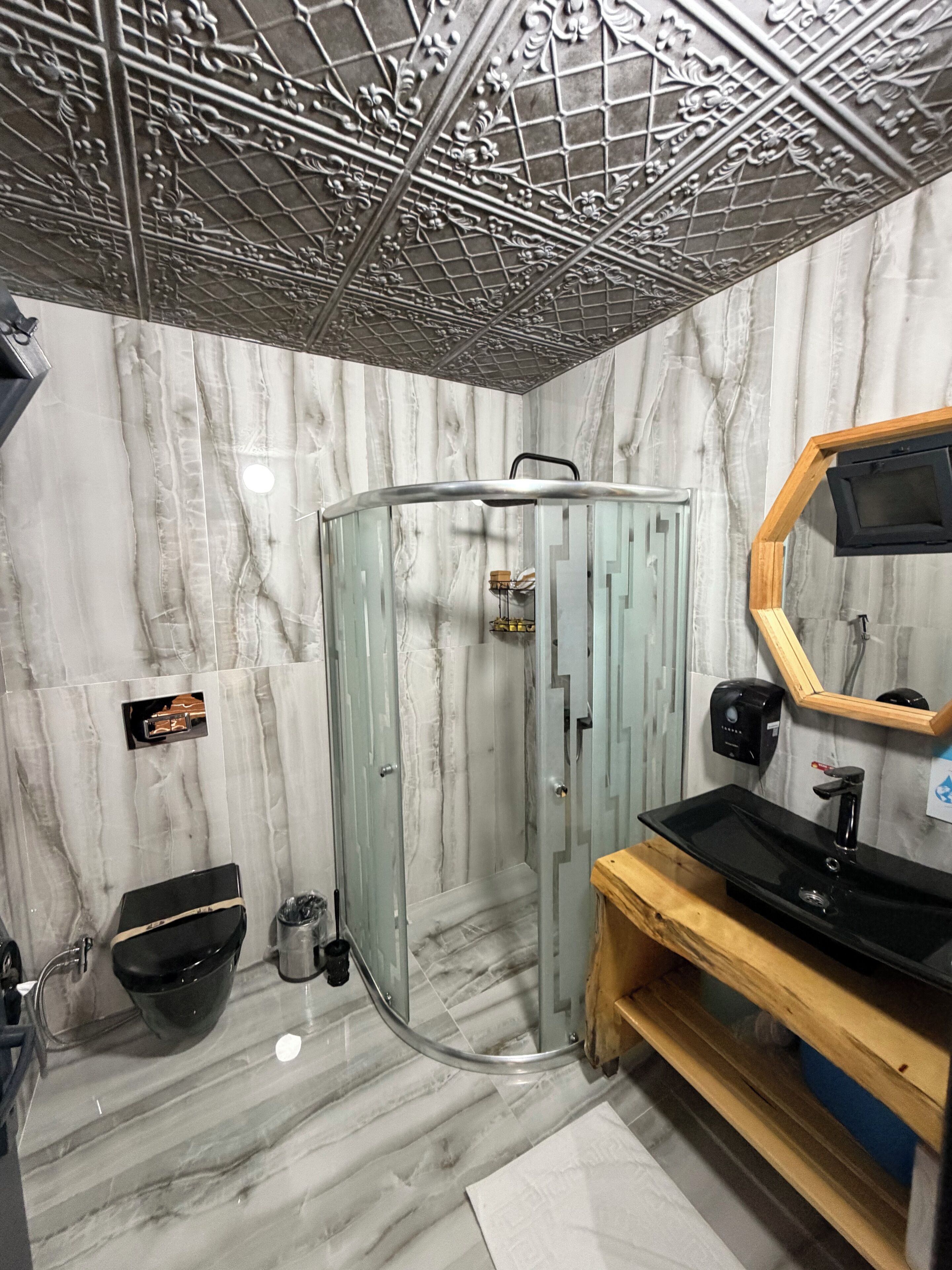 Une salle de bain avec un mur en marbre, une douche en verre, un meuble-lavabo en bois avec un évier noir et un miroir avec un cadre en bois.