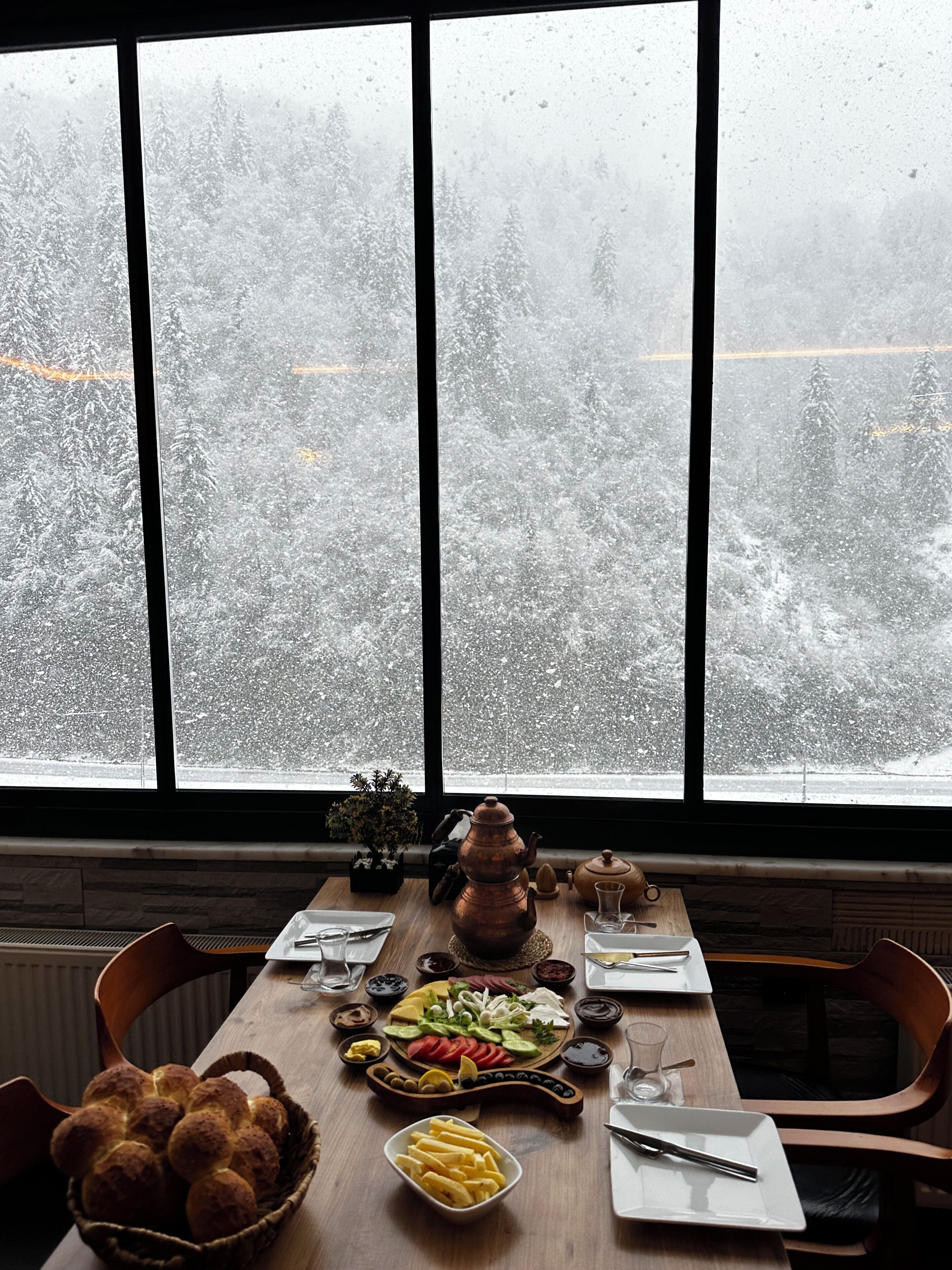 Una mesa de comedor con diversos alimentos y una tetera, con vistas a un bosque nevado a través de una ventana.