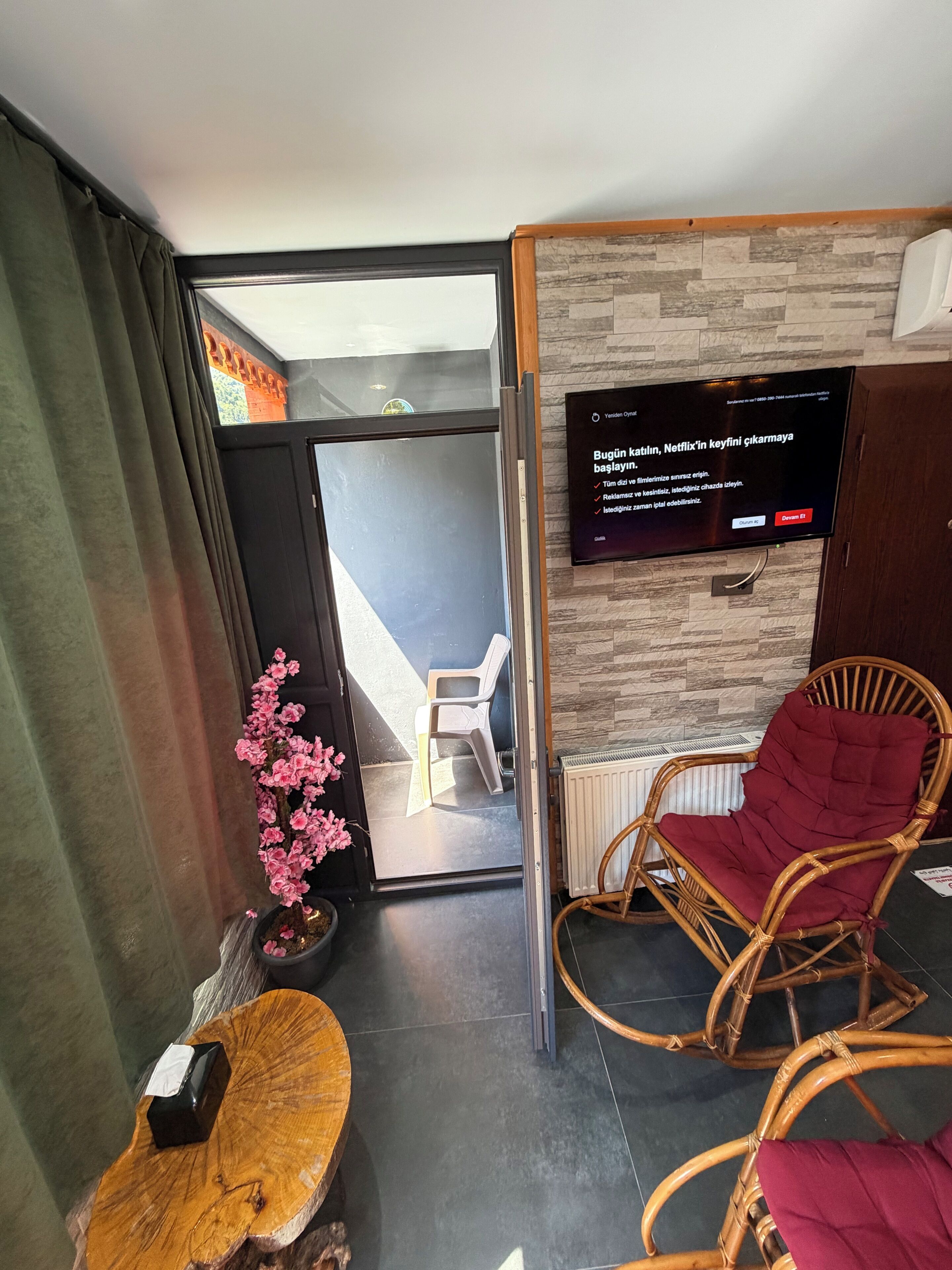 Una habitación moderna con una puerta corrediza de vidrio que da a un balcón, un televisor de pared, una silla de mimbre y una pequeña mesa de madera.