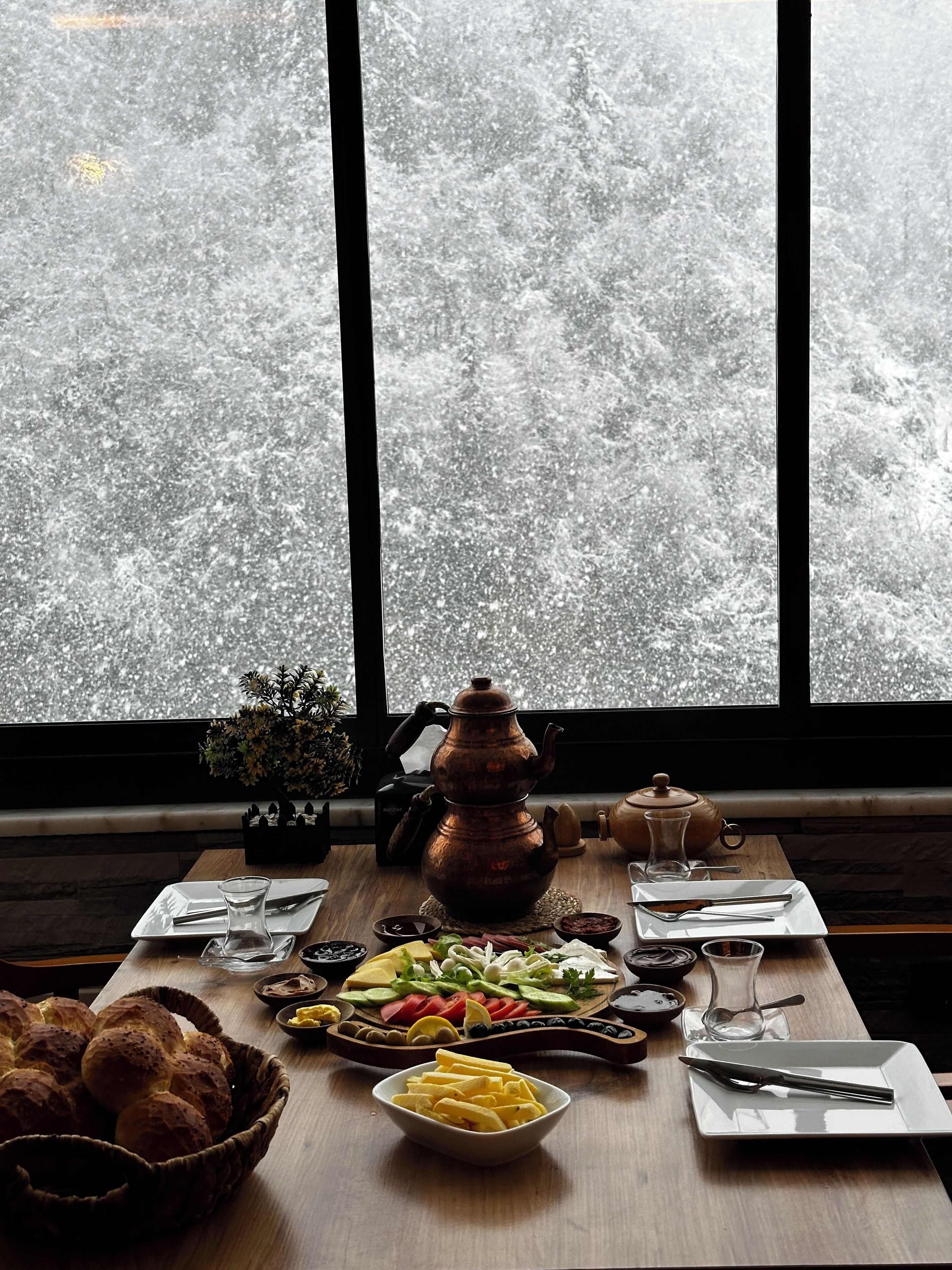 A dining table set with various dishes, a teapot, and a view of a snowy landscape through large windows.