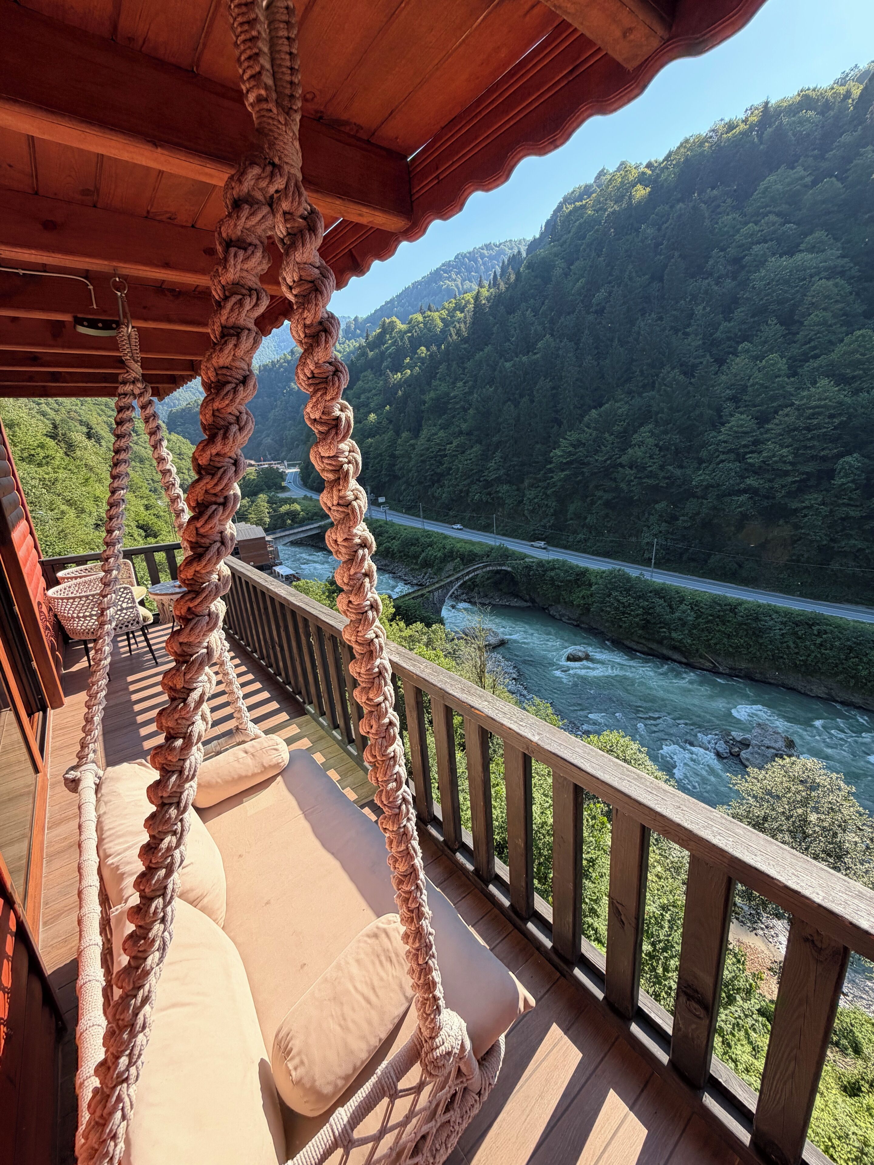 A wooden deck with a rope swing, a cushioned seating area, and a view of a river and forested hills.