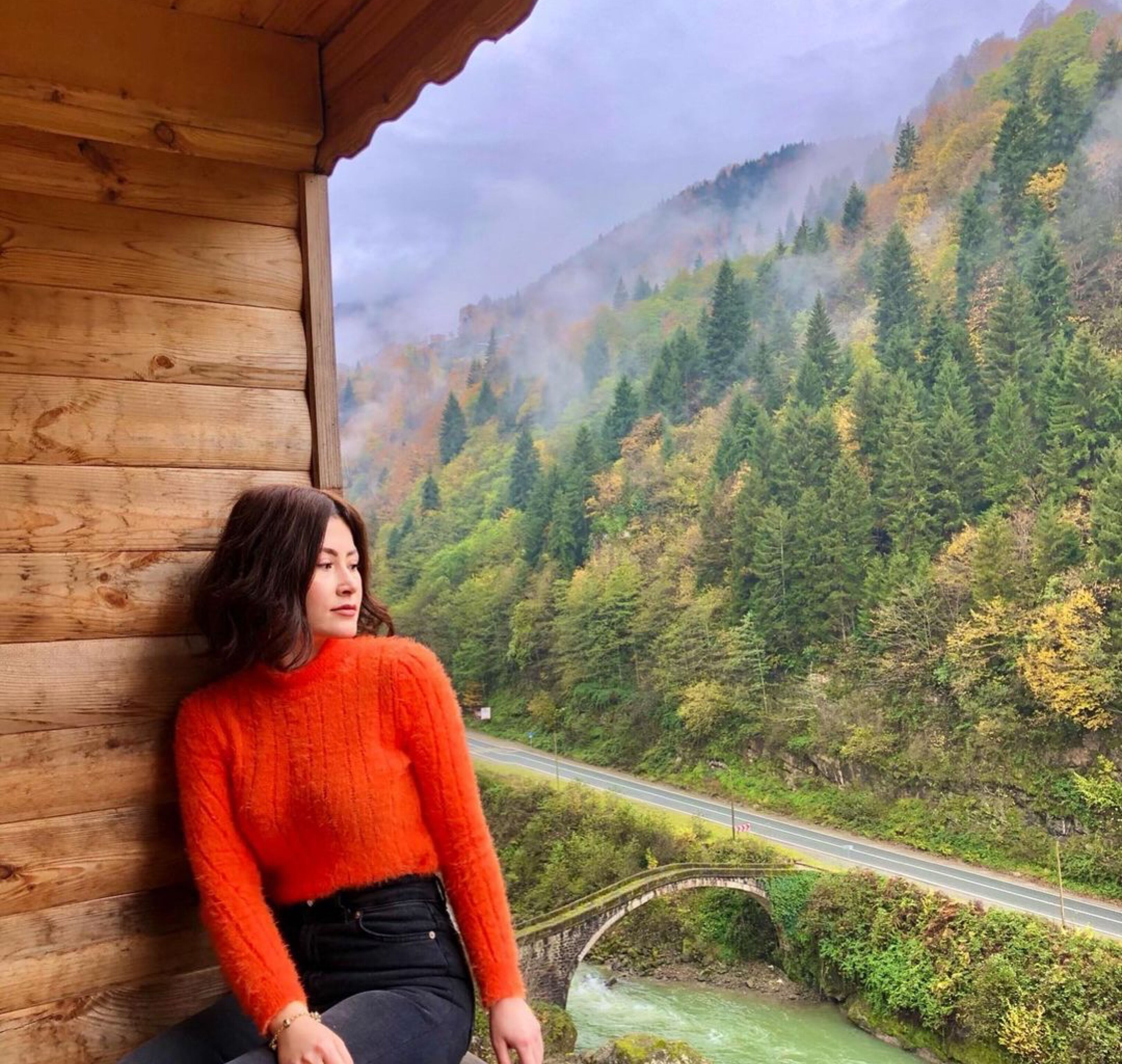 A person sitting by a wooden cabin with a view of a forested mountain and a bridge over a river.
